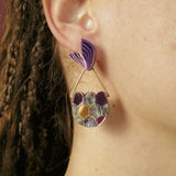 A close-up of a person’s ear wearing a large, artistic earring that channels whimsical elegance with its purple leaf-shaped top and circular lower section. Perfect for mermaid lovers, this accessory complements their brown braided hair beautifully.