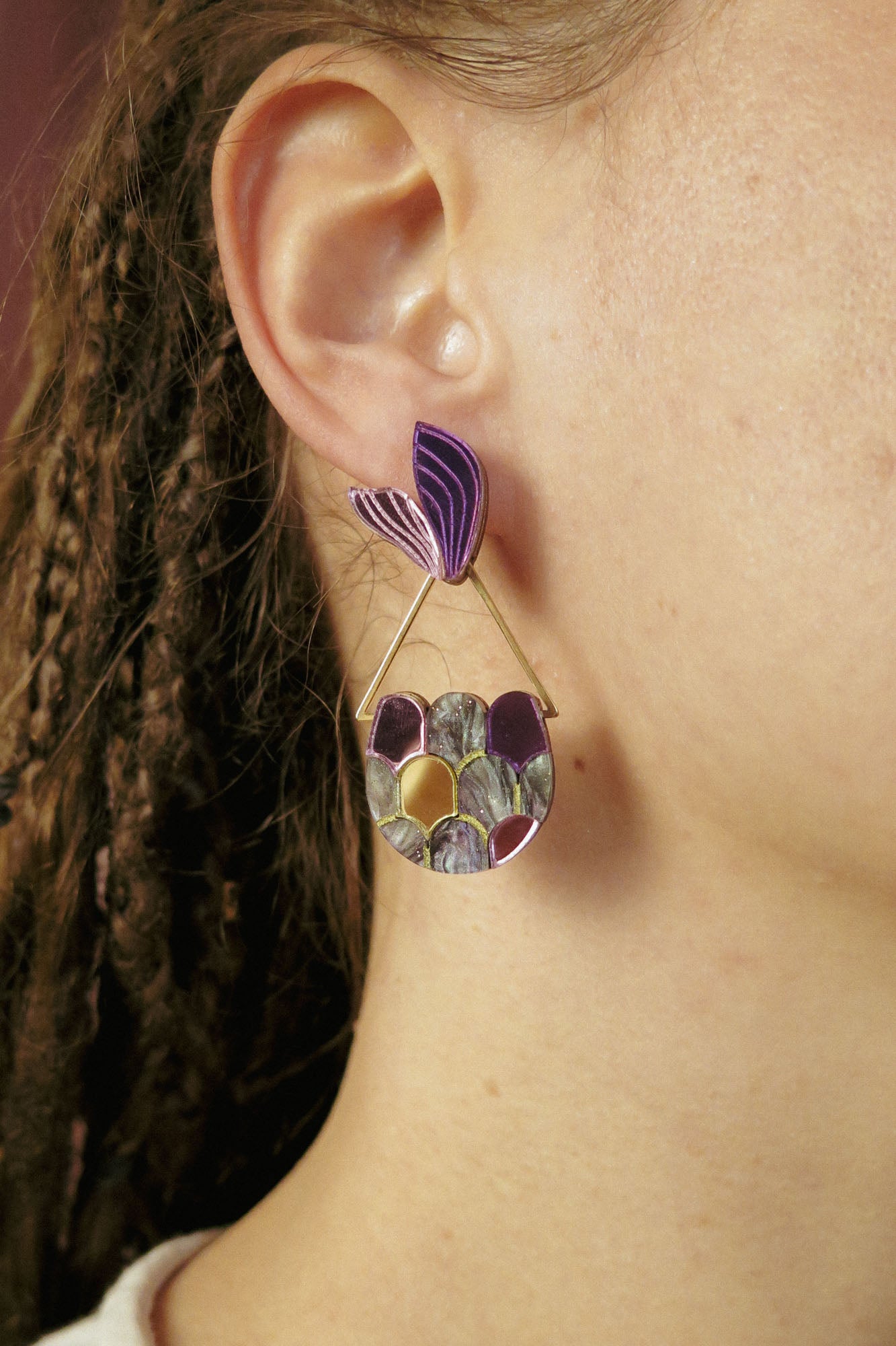 A close-up of a person’s ear wearing a large, artistic earring that channels whimsical elegance with its purple leaf-shaped top and circular lower section. Perfect for mermaid lovers, this accessory complements their brown braided hair beautifully.