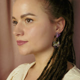 A woman with long brown dreadlocks and natural makeup looks to the side, embodying whimsical elegance. She wears a cream-colored top and geometric Mermaid Tail Earrings with purple accents. The background is softly blurred with pinkish curtains.