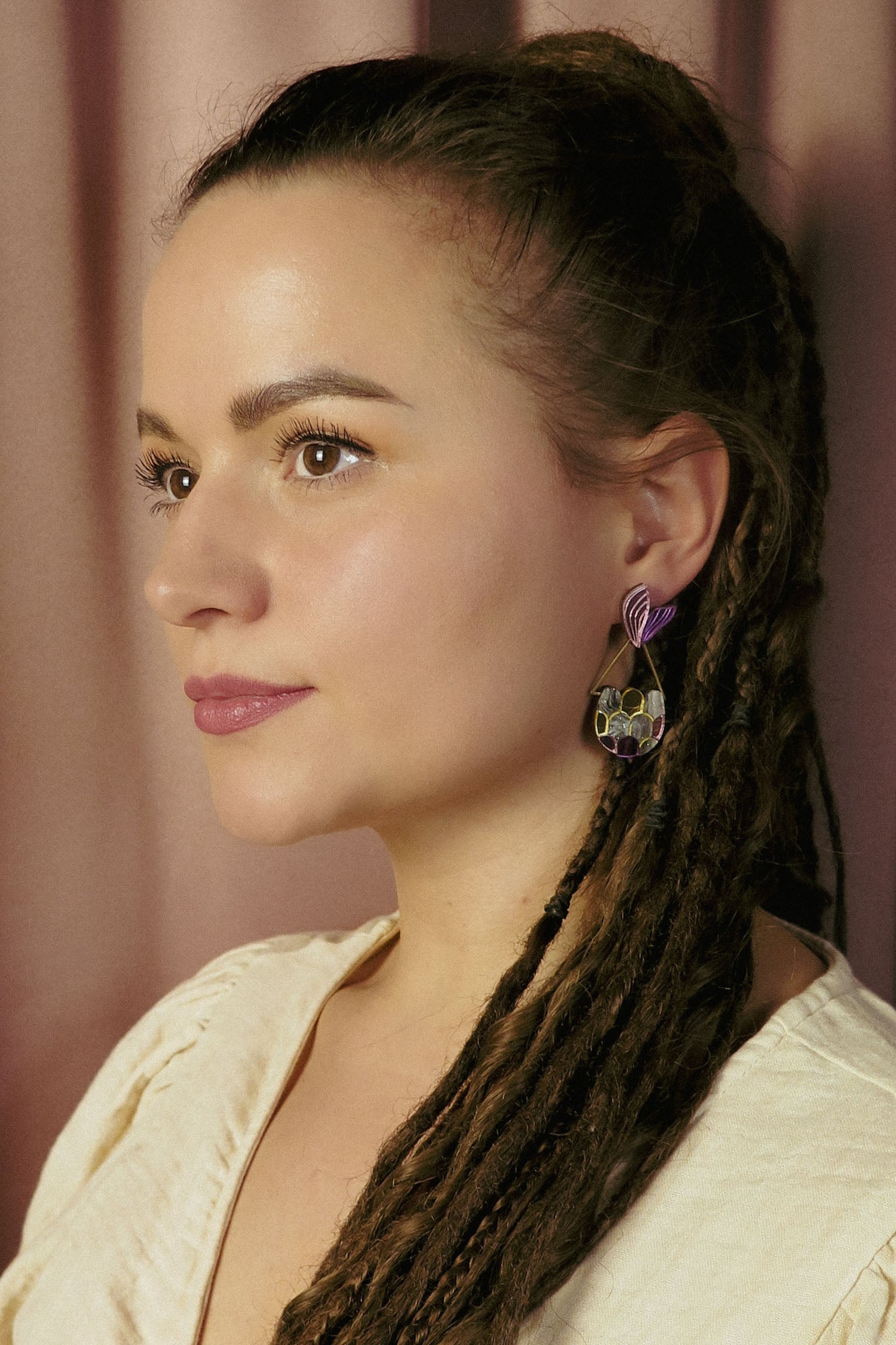 A woman with long brown dreadlocks and natural makeup looks to the side, embodying whimsical elegance. She wears a cream-colored top and geometric Mermaid Tail Earrings with purple accents. The background is softly blurred with pinkish curtains.