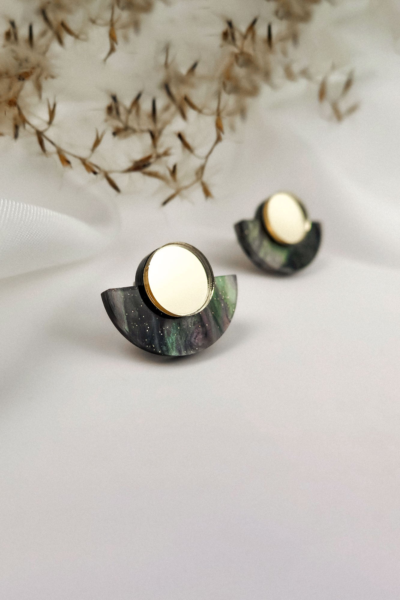 A pair of minimalist geometric stud earrings featuring a gold mirrored circle atop a dark, semi-circular marbled base, displayed on a white surface with dried plant accents in the background.