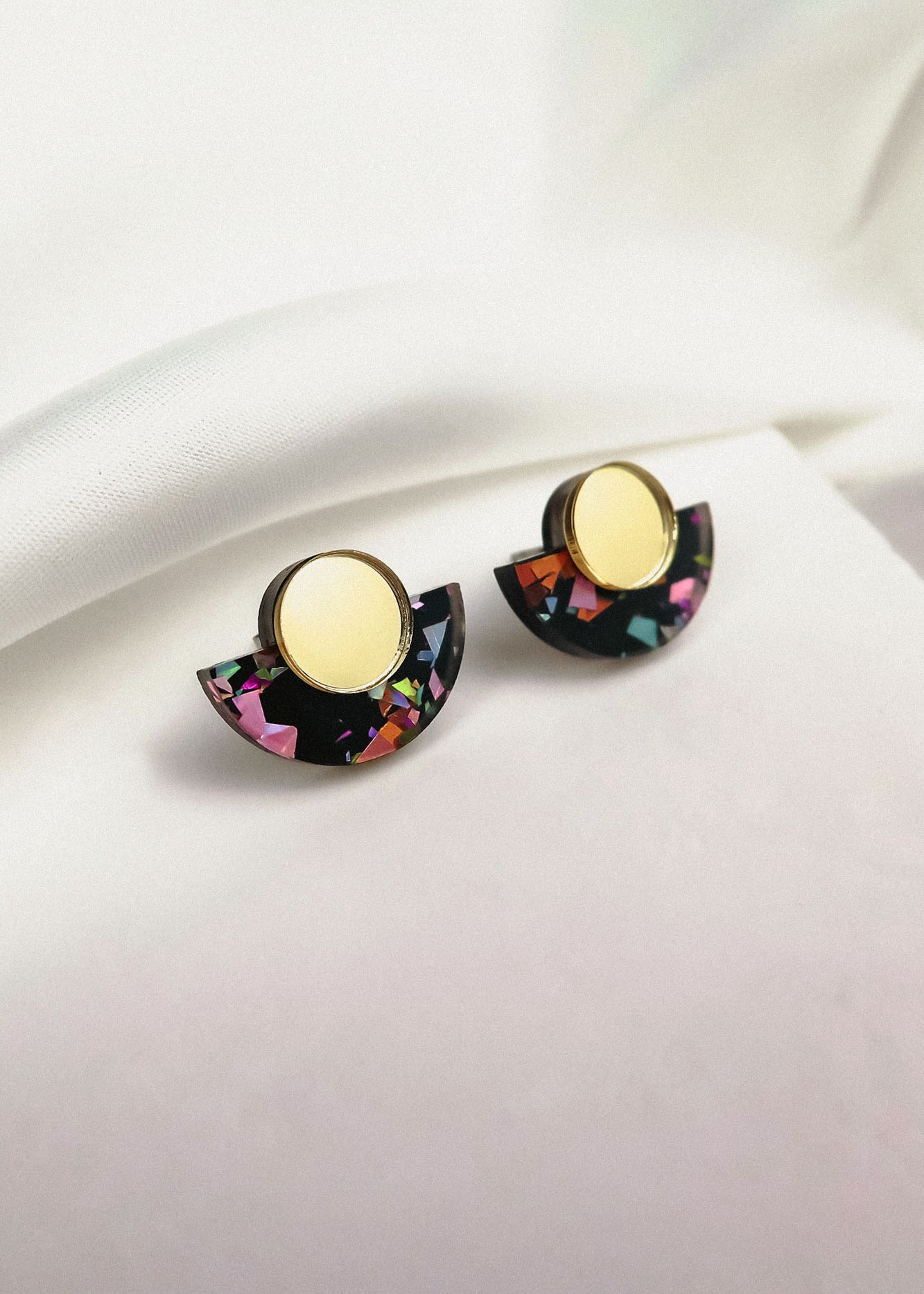 Minimalist Geometric Stud Earrings featuring gold circular tops and black semicircular bottoms with colorful, confetti-like patterns. These lightweight, hypoallergenic earrings are displayed on a white fabric background.
