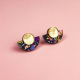 A pair of minimalist geometric stud earrings with gold circular tops and black semicircular bases featuring vibrant, multicolored confetti-like patterns, these hypoallergenic, lightweight pieces are displayed on a pink background.