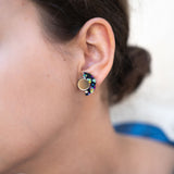 Close-up of a woman’s ear wearing colorful, crescent-shaped Minimalist Geometric Stud Earrings with a round gold stud. The womans dark hair is tied back in a bun. These hypoallergenic earrings are lightweight and set against a softly blurred background.