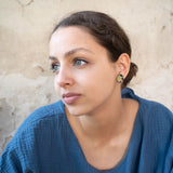 A young woman with light eyes and brown hair pulled back, wearing a textured blue top and lightweight Minimalist Geometric Stud Earrings, looks thoughtfully to the side against a beige, textured wall.