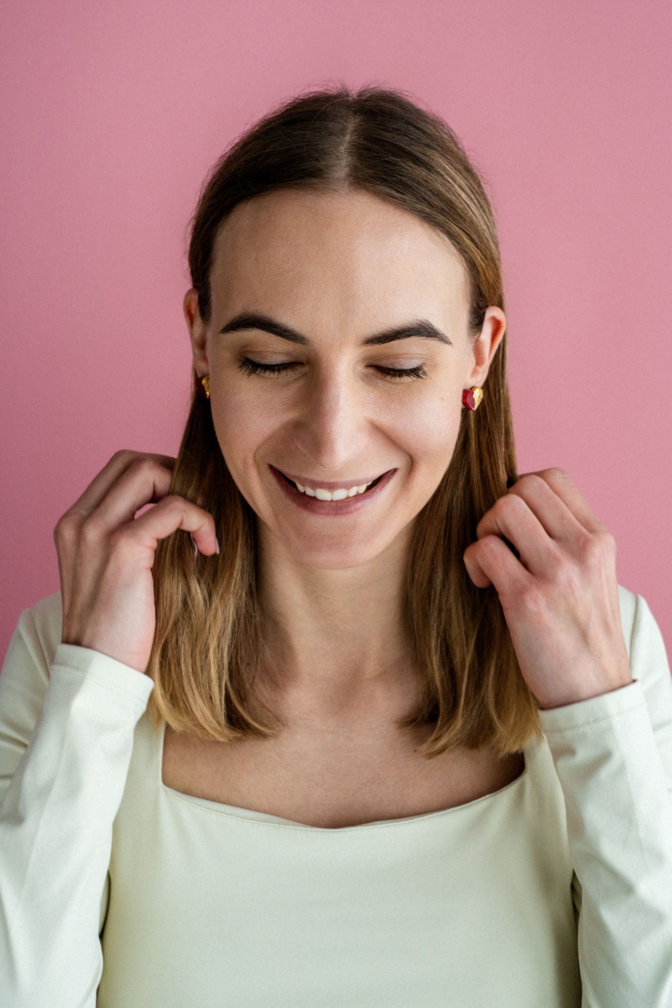 A woman with shoulder-length brown hair and a white top smiles with her eyes closed against a pink background, touching her hair with both hands, while wearing delicate handcrafted earrings.