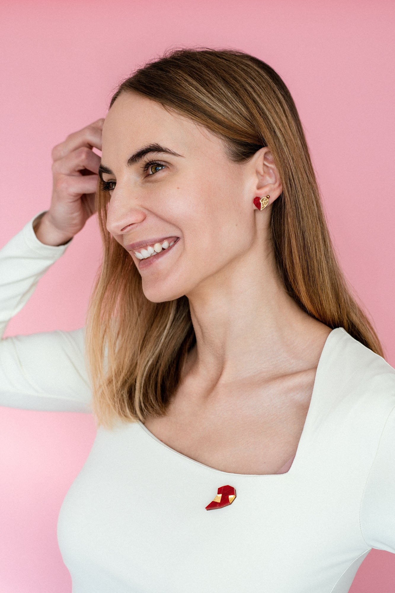 A smiling woman with straight, light brown hair wears a white, square-neck top and romantic jewelry—handcrafted earrings and a matching pin in red-and-gold geometric shapes—against a pink background. She raises her left hand to her hair.