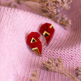 A pair of handcrafted geometric red and gold earrings shaped like hearts rest on textured pink fabric, with small dried flowers placed nearby—a perfect romantic gift.