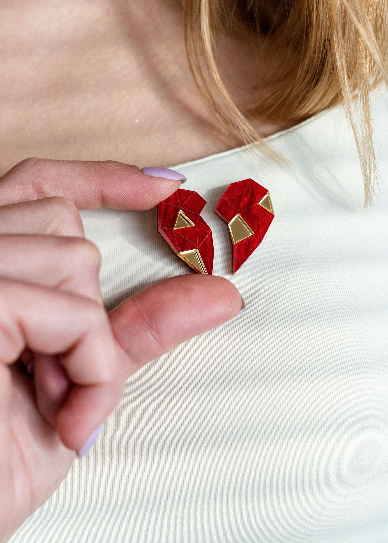 A person wearing a white top holds a red, geometric, handcrafted One Love Halves Pin against their chest. The heart-shaped pin is split into two pieces with gold accents, making it a meaningful romantic gift.