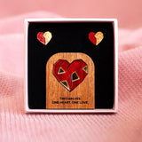 A jewelry set with geometric polygon hearts earrings and a matching pin displayed in a box. The pin features a red and gold heart on wood with the text: TWO HALVES. ONE HEART. ONE LOVE.—the perfect Valentine’s statement. Soft pink fabric background.