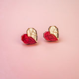 Two origami hearts, each half red and half gold, are placed side by side on a smooth, light pink background, resembling handcrafted earrings or romantic jewelry.