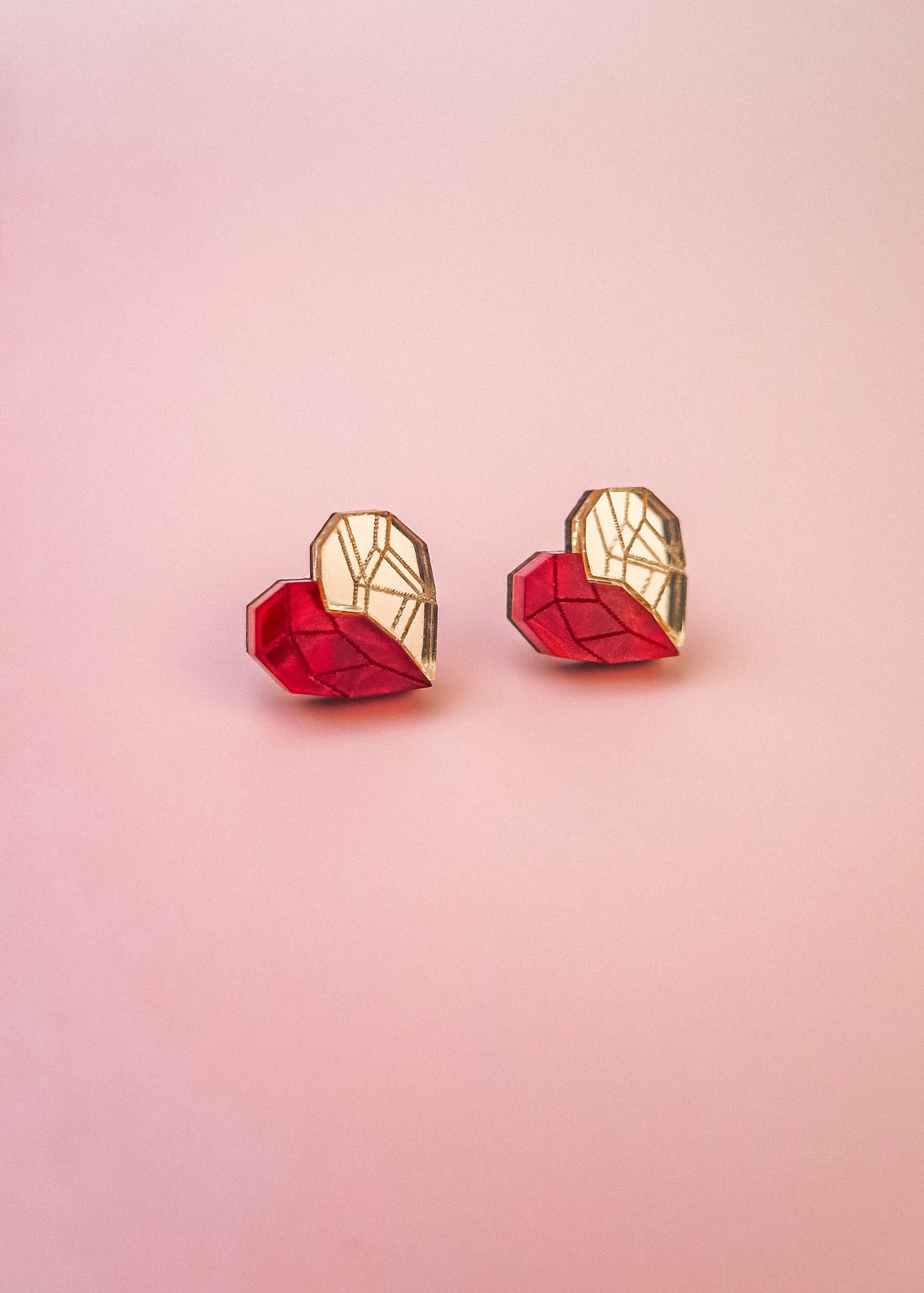 Two origami hearts, each half red and half gold, are placed side by side on a smooth, light pink background, resembling handcrafted earrings or romantic jewelry.