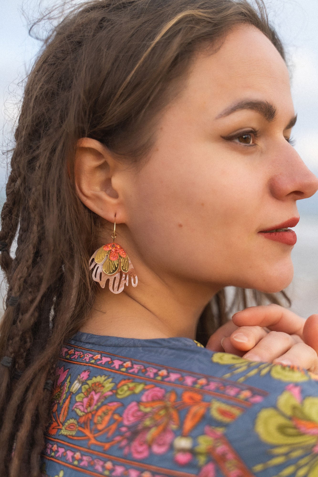 A woman with brown dreadlocks and winged eyeliner looks to the side, wearing a colorful floral-patterned garment and striking Organic Floral Hoops with pink and gold accents. She has a slight smile, her hands gently clasped.