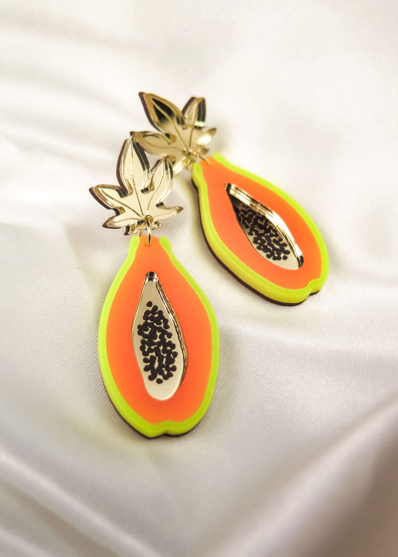 A pair of colorful Papaya Earrings shaped like papaya slices, crafted from neon acrylic glass with gold leaf tops, orange and yellow accents, black seed details, and allergy-free stainless steel posts, displayed on white satin fabric.