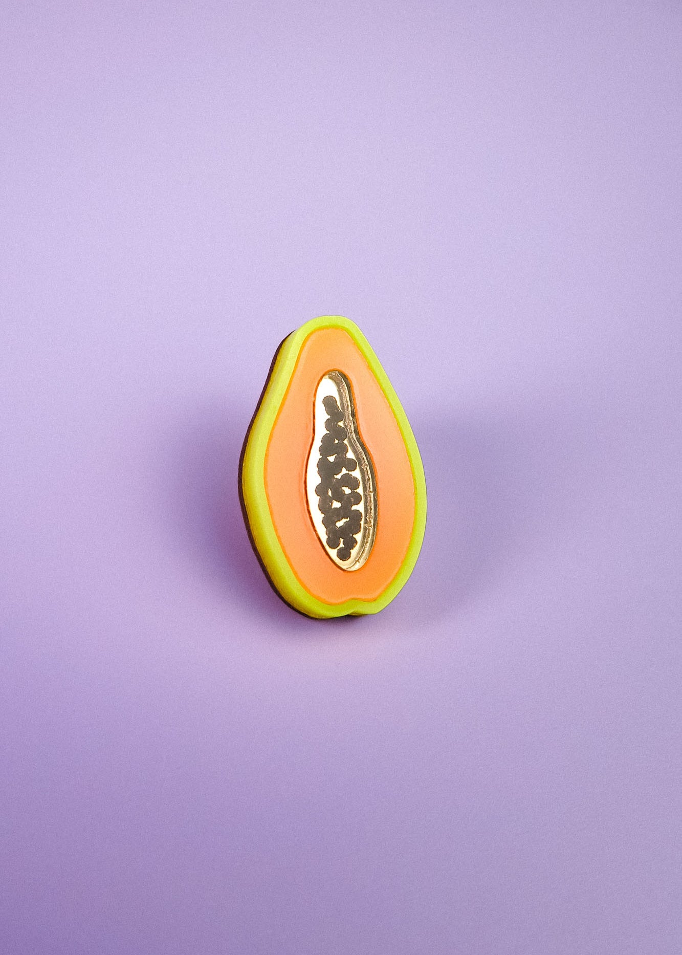 A handcrafted cookie decorated to look like a sliced papaya, with neon orange, yellow, and green icing plus black seeds, stands upright on a purple surface with a purple background—just like a Papaya Pin come to life.