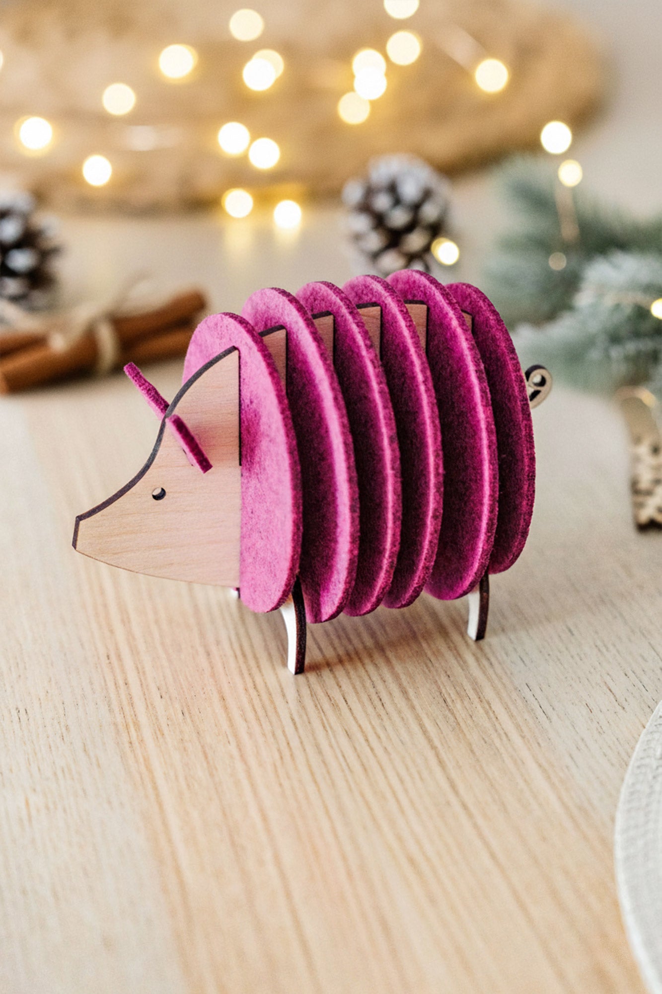 A decorative hedgehog figurine, crafted from wood and vibrant purple felt discs, sits on a light wooden table—an ideal housewarming gift—surrounded by festive lights, pinecones, and cinnamon sticks blurred in the background.