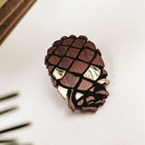 A brown, pinecone-shaped brooch crafted from recycled wood with shiny accents sits on a white surface, surrounded by blurred green leaves and a wooden object in the background.