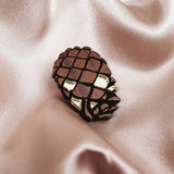 A gold and brown Pinecone Brooch, crafted from sustainable recycled wood, rests on shiny, light beige satin fabric with soft folds.