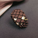A sustainable pinecone brooch made from recycled wood with gold accents lies on a textured dark surface near a pink satin fabric.