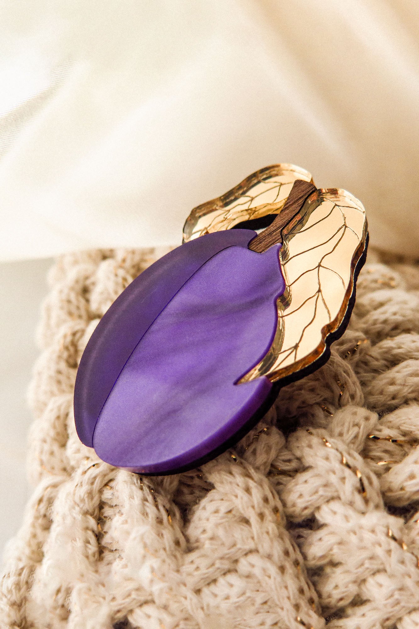 A purple brooch shaped like a plum with gold-colored leaves, handmade from recycled wood, rests on a knitted beige fabric.