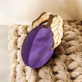 A handmade purple plum-shaped brooch with two gold and ivory leaves is attached to a chunky beige knit fabric. Cream-colored fabric appears in the background.