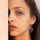 A woman with long brown hair and elegant purple plum earrings looks directly at the camera, standing next to a textured beige wall, with soft natural light highlighting her face.