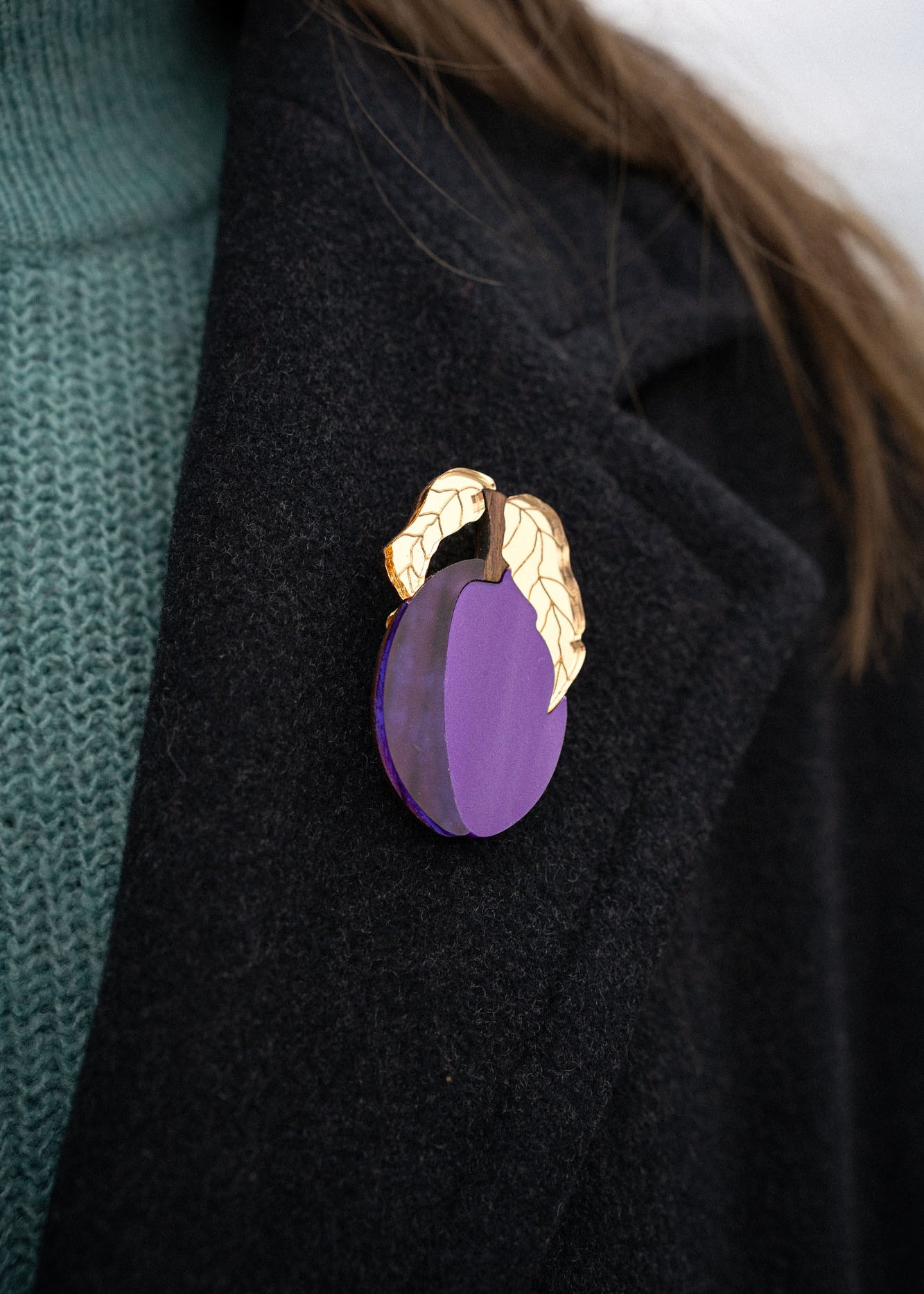 A person wearing a dark coat with an elegant brooch shaped like a purple fruit and two gold-colored leaves pinned to the lapel. The person is also wearing a teal knit sweater.