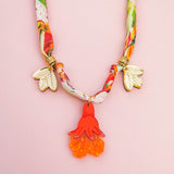 A colorful handmade necklace with a floral-patterned fabric band, two gold flower charms, and a central pendant shaped like an orange and red flower—perfect as a unique pomegranate flower necklace—displayed on a light pink background.