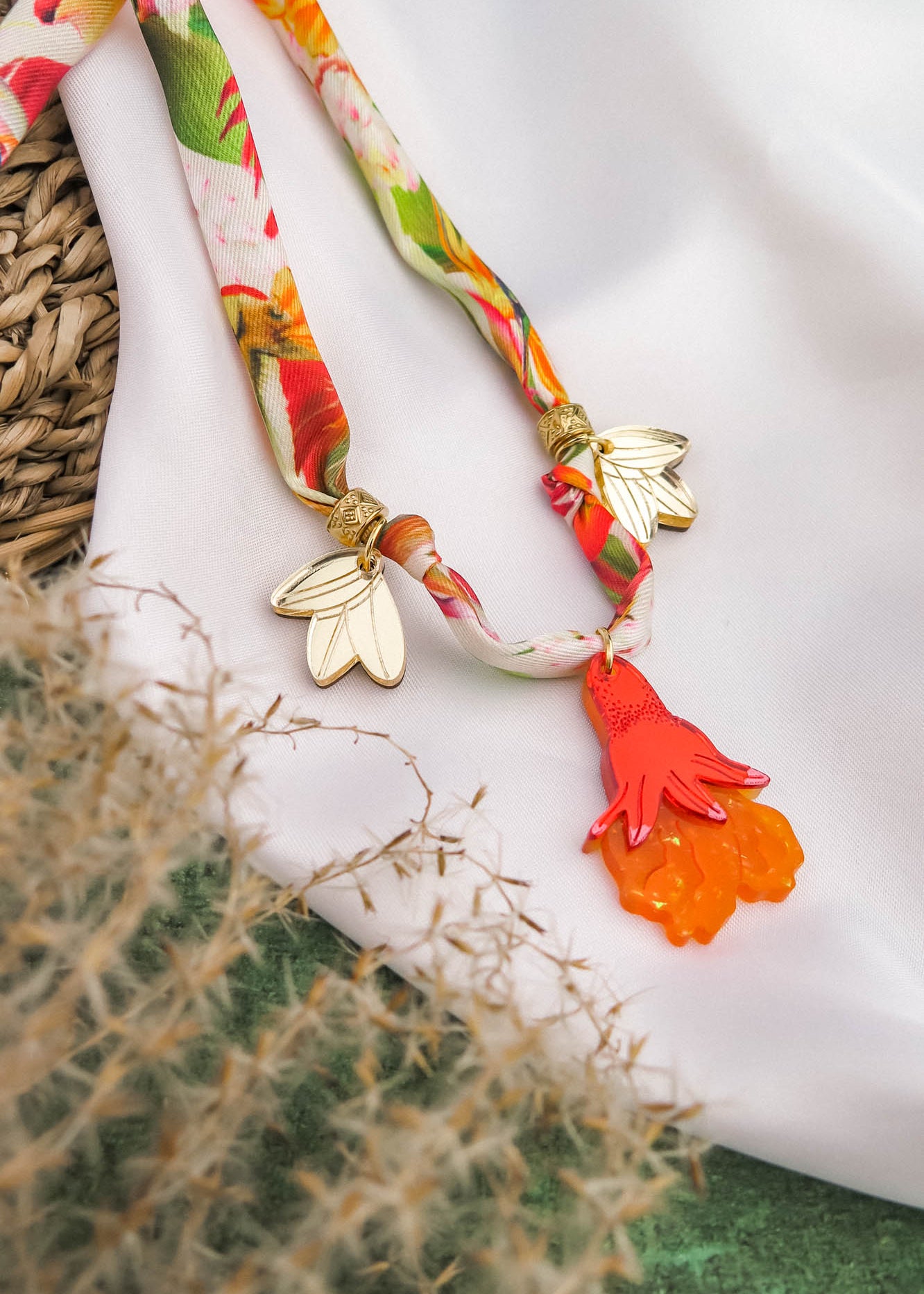 A colorful silk cord necklace with gold leaf charms and an orange-red hand-shaped pendant, displayed on white fabric with a woven basket and beige dried grass nearby.