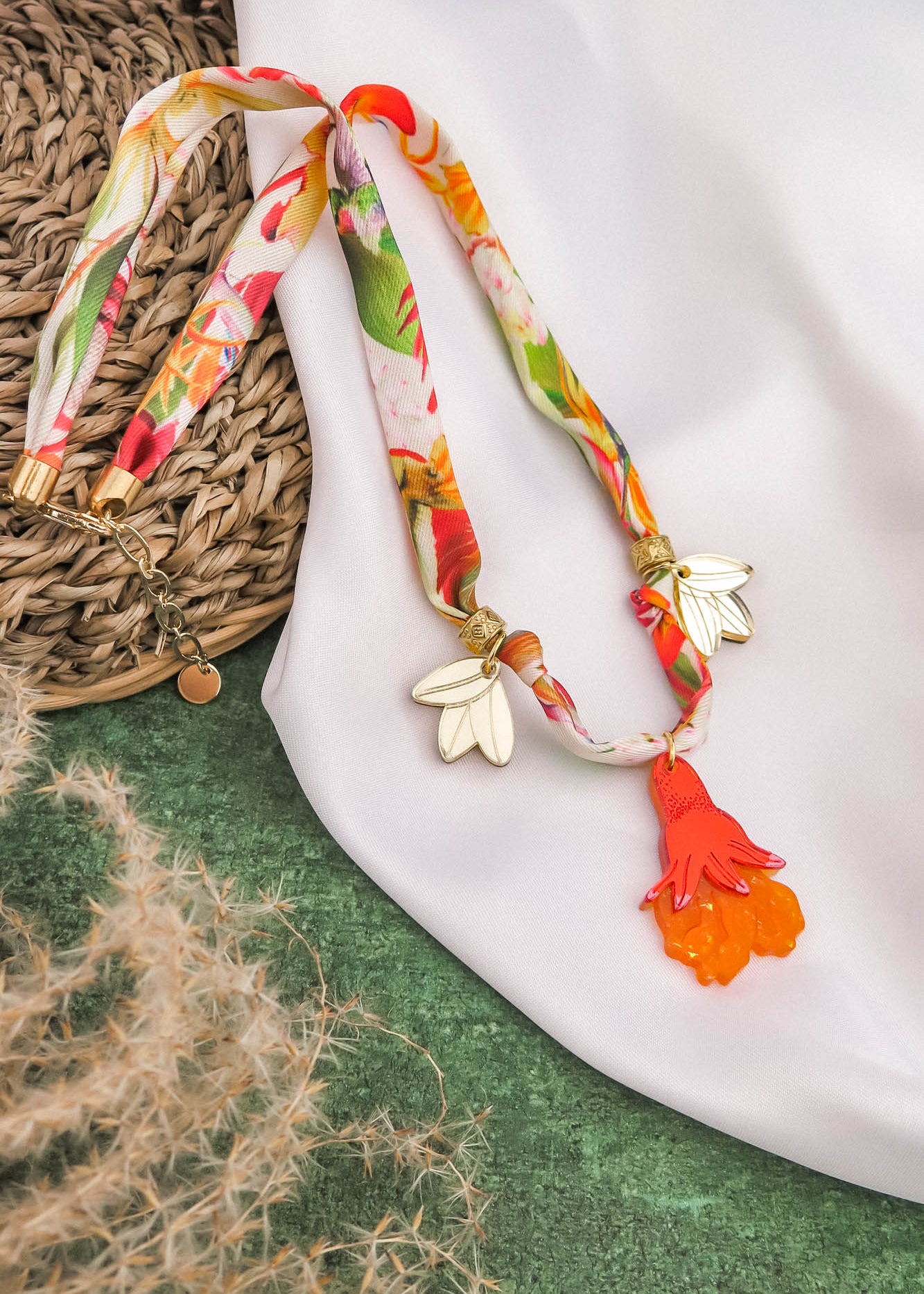 A colorful handmade necklace with floral patterns, featuring two gold and white wing-shaped charms and a bright orange hand-shaped pendant, is displayed on a white cloth and green textured background.