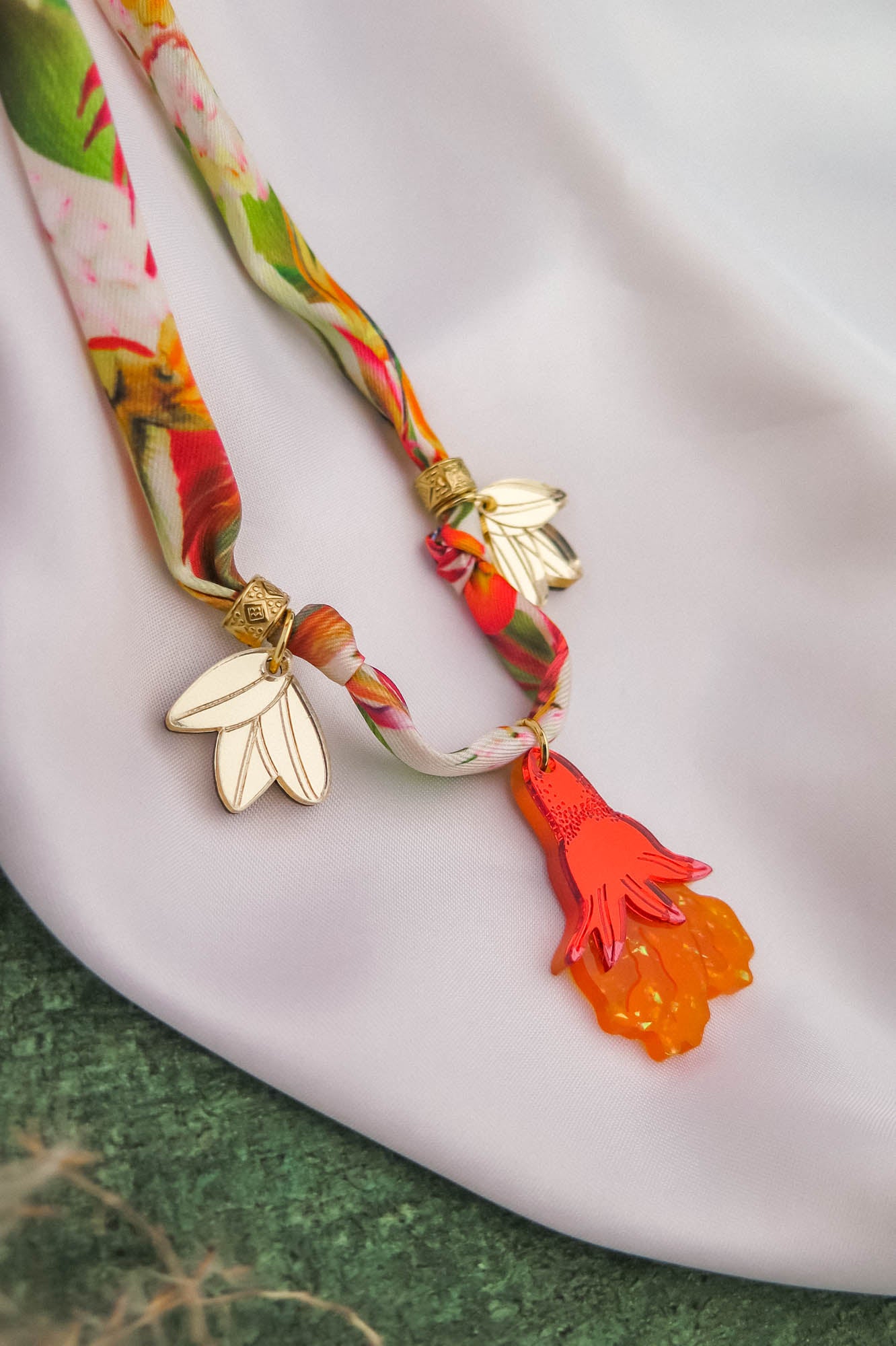 A colorful floral scarf with gold and white flower-shaped clips and a bright orange and pink pendant, displayed on a white fabric surface with a green background, pairs beautifully with a handmade pomegranate flower necklace.