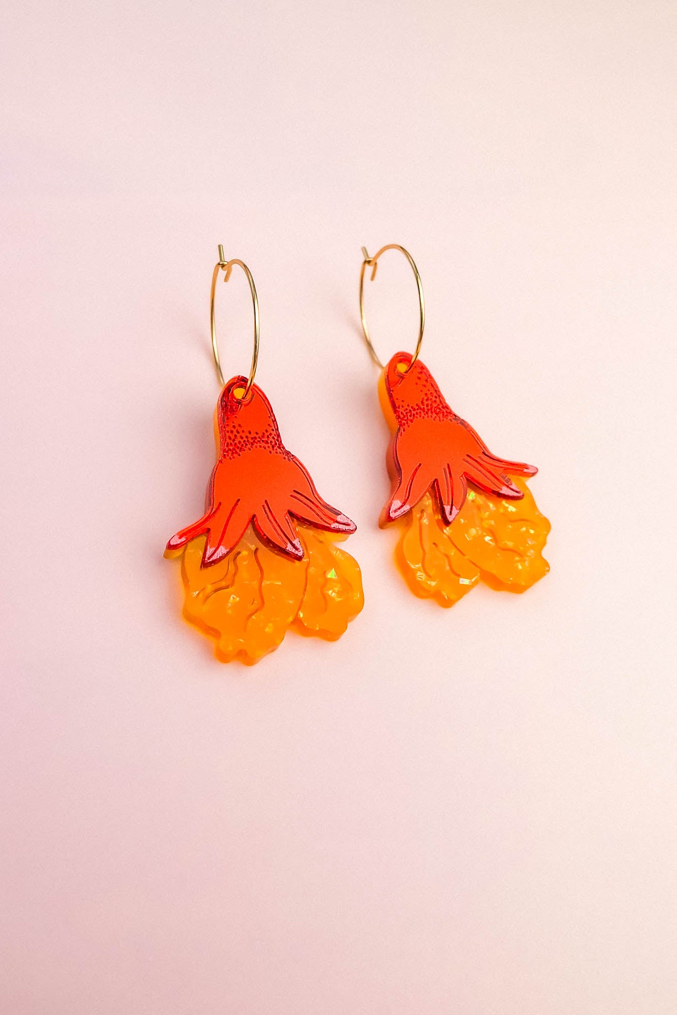 A pair of Little Turtle Hoop Earrings featuring gold hoops and pendants shaped like orange and yellow hand-drawn flowers, set against a pale pink background—beautifully handcrafted earrings for a playful, artistic touch.