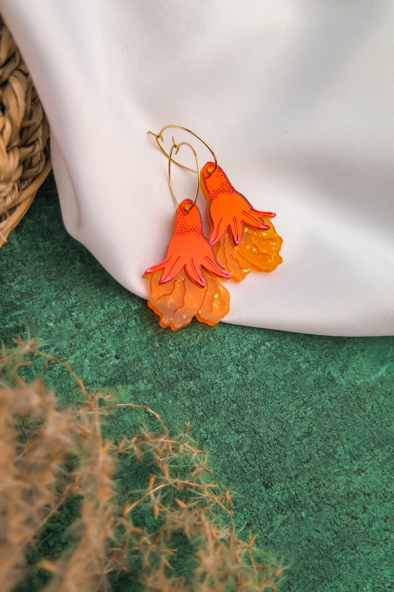 A pair of orange and yellow handcrafted earrings, shaped like stylized flowers or flames, rest on a white fabric backdrop. Green textured ground and part of a woven basket are visible, highlighting these unique gold hoops.