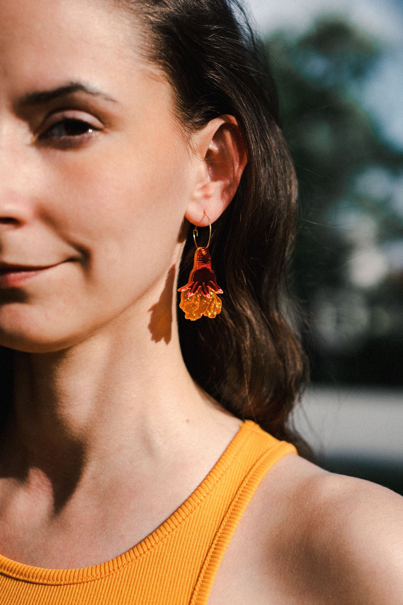 A woman with long brown hair wears handmade floral earrings and a sleeveless yellow top, standing outdoors in natural light. Only the left side of her face and shoulder are visible.