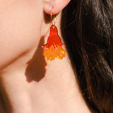 Close-up of a person’s ear wearing bold, handmade floral earrings shaped like a red and yellow jellyfish. The person has long brown hair and stands in bright sunlight, adding summer vibes to these unique accessories.