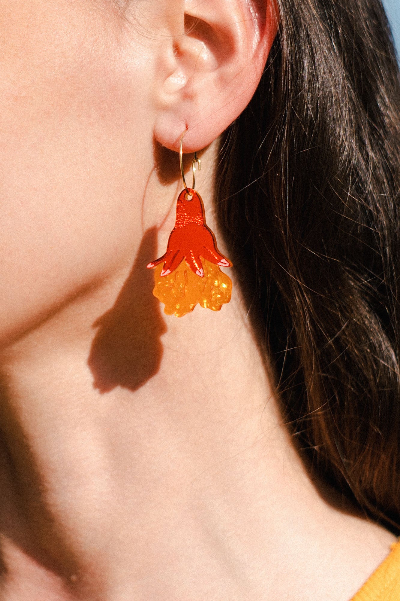 Close-up of a person’s ear wearing bold, handmade floral earrings shaped like a red and yellow jellyfish. The person has long brown hair and stands in bright sunlight, adding summer vibes to these unique accessories.