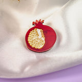 A handcrafted Pomegranate Pin, shaped like a red pomegranate with gold detailing, rests on white satin fabric. Some dried plants are visible in the background and a purple surface appears at the bottom—an elegant fashion accessory.