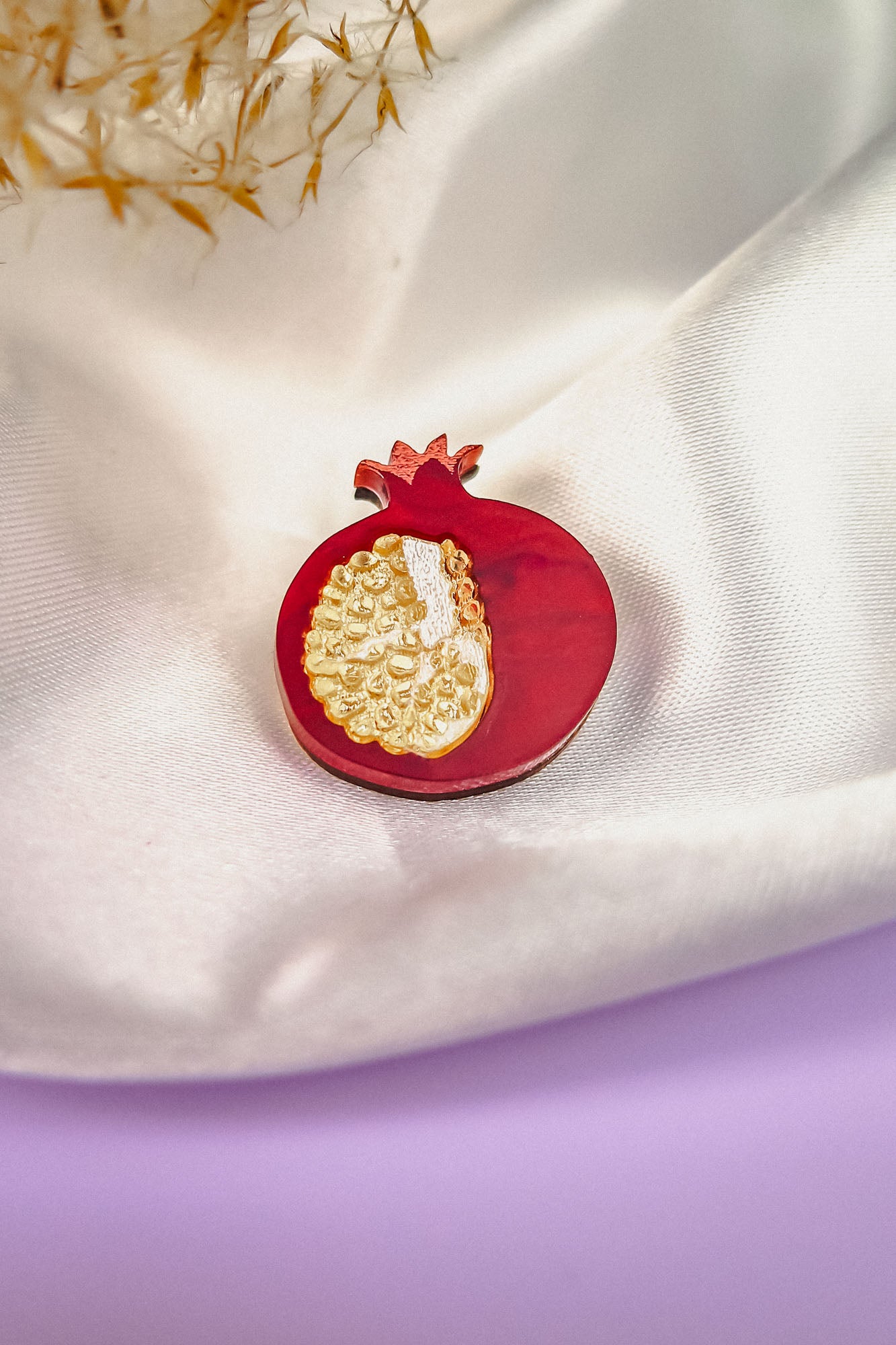 A handcrafted Pomegranate Pin, shaped like a red pomegranate with gold detailing, rests on white satin fabric. Some dried plants are visible in the background and a purple surface appears at the bottom—an elegant fashion accessory.