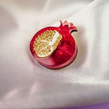 A handcrafted Pomegranate Pin brooch with red and gold details rests on a white satin fabric background, making it an elegant fashion accessory.