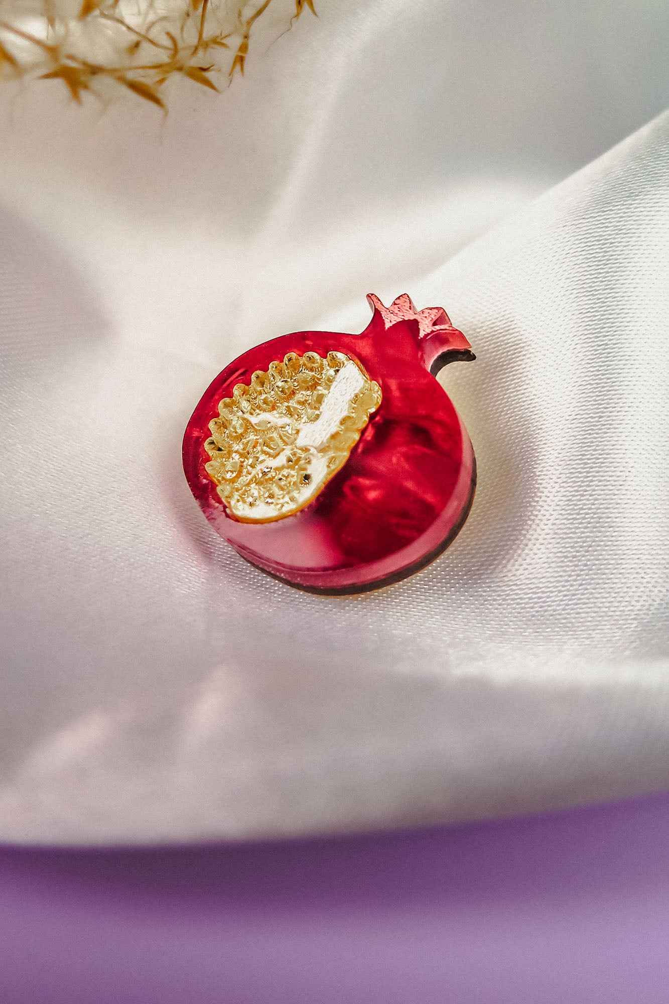 A handcrafted Pomegranate Pin brooch with red and gold details rests on a white satin fabric background, making it an elegant fashion accessory.