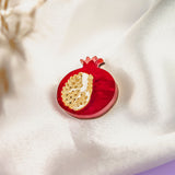 A handcrafted red pomegranate-shaped brooch with gold detailing rests on smooth, white satin fabric, making this Pomegranate Pin a unique fashion accessory. A blurred floral element decorates the upper left corner.