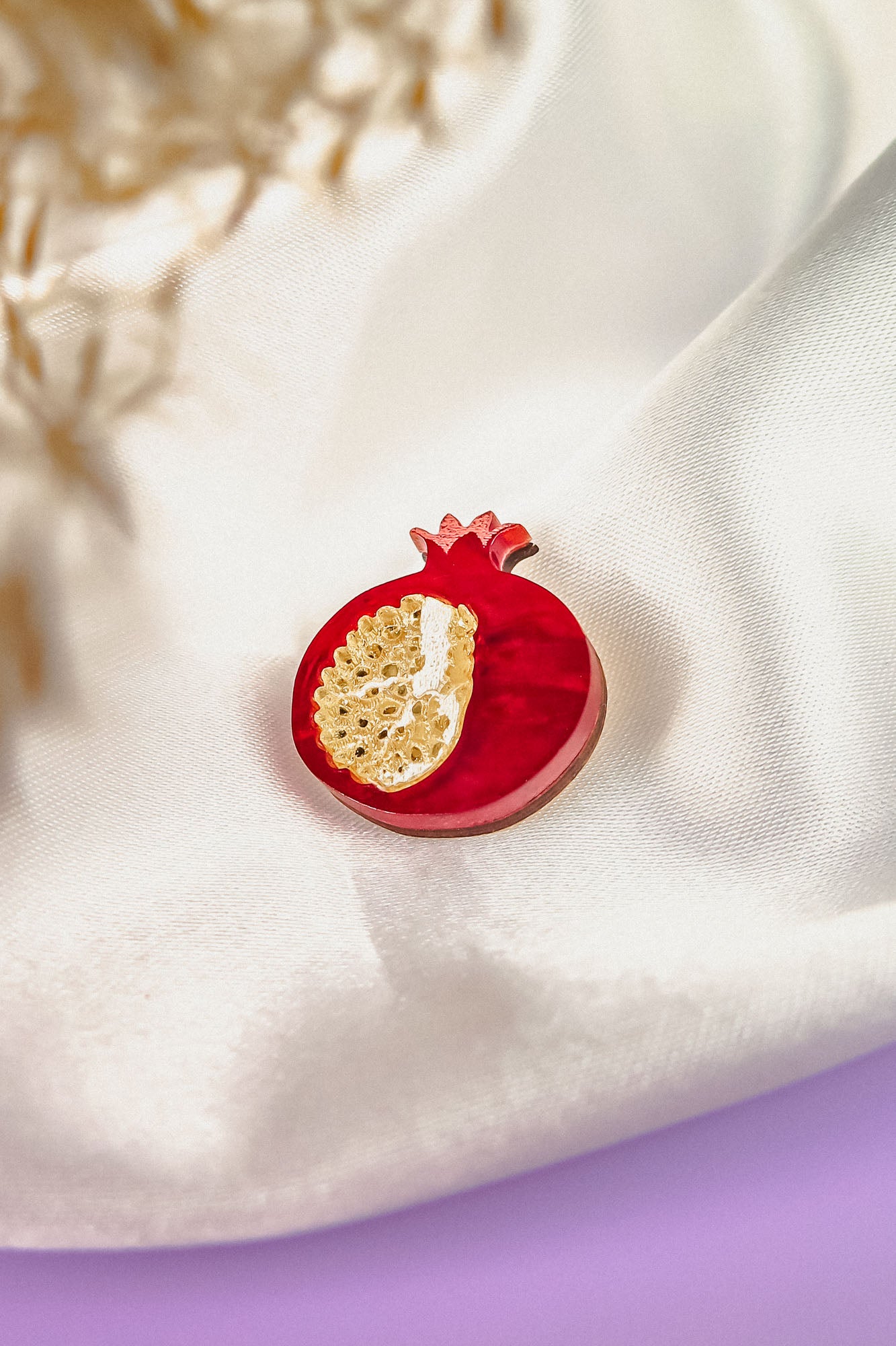 A handcrafted red pomegranate-shaped brooch with gold detailing rests on smooth, white satin fabric, making this Pomegranate Pin a unique fashion accessory. A blurred floral element decorates the upper left corner.