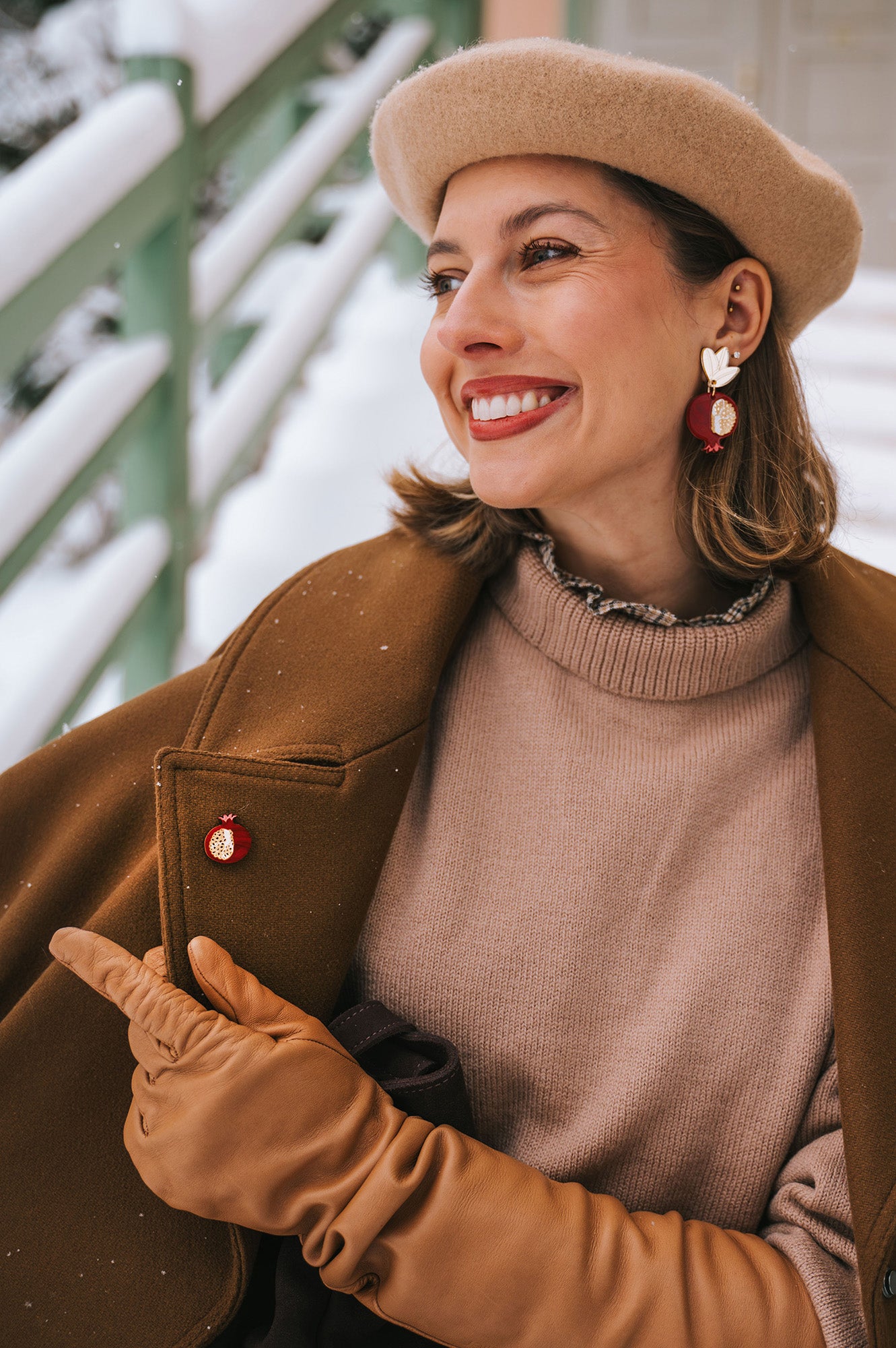 A smiling woman wearing a beige beret, brown coat, tan gloves, and statement earrings with red and gold accents sits outside in a snowy setting, pointing to a handcrafted Pomegranate Pin—a unique fashion accessory on her coat.