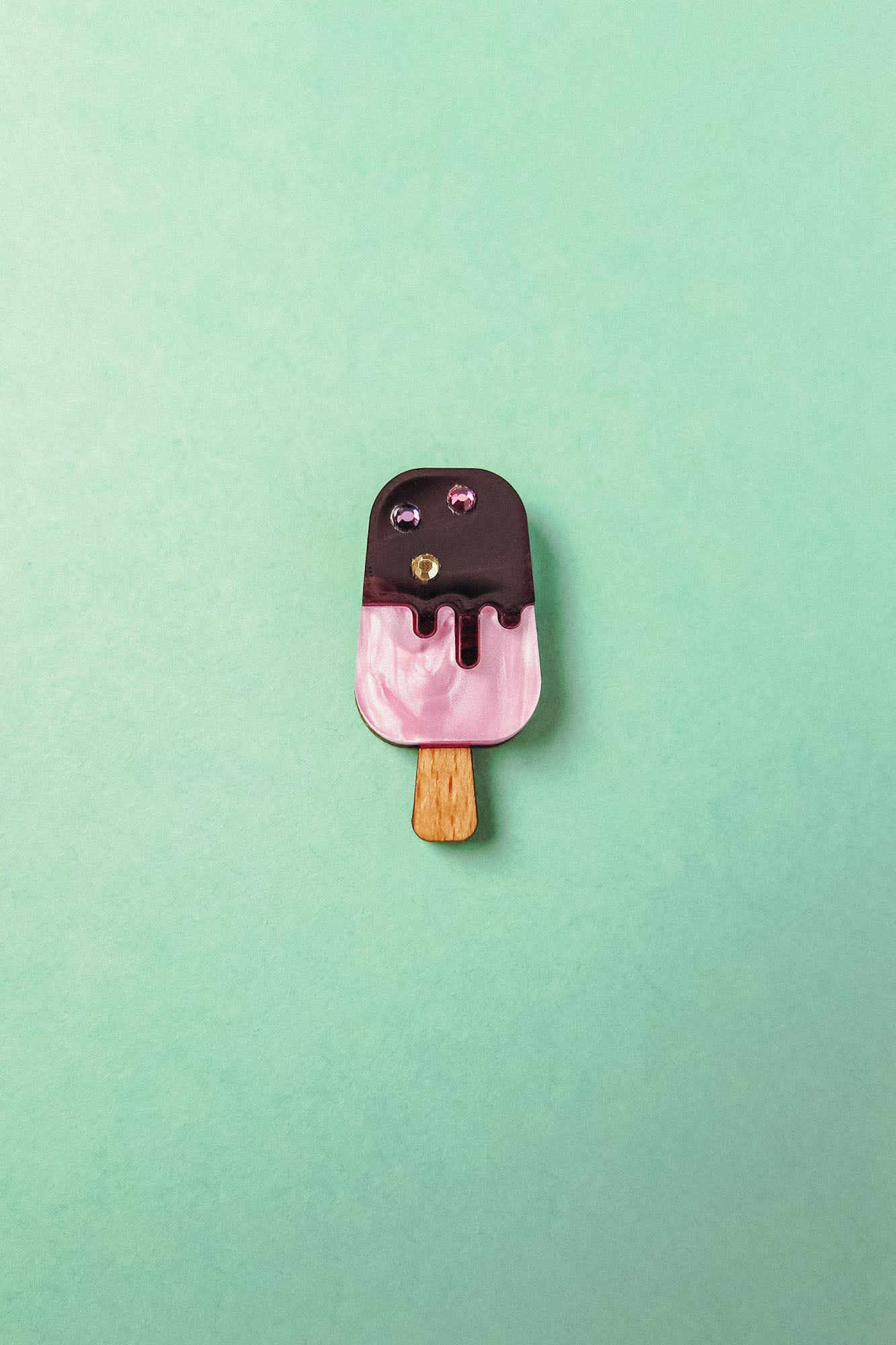 A small, pink and brown handcrafted popsicle pin with glittery gem accents and rhinestones, placed on a mint green background.