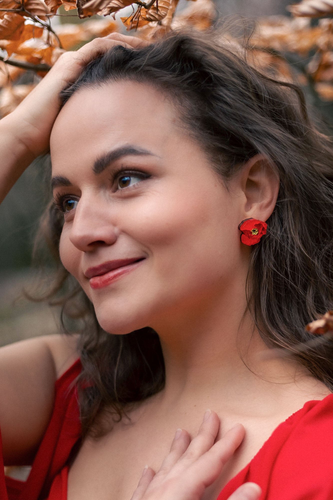 A woman with wavy brown hair, wearing a red dress and lightweight acrylic earrings with a poppy design, stands outdoors among autumn leaves, smiling softly with one hand on her head and the other on her chest.