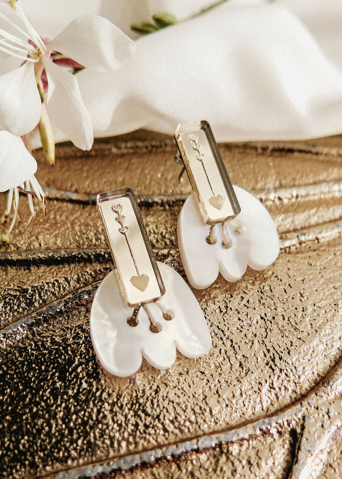 Rectangular gold earrings with engraved key and heart designs rest on white shell-like bases, displayed on a textured metallic surface with white flowers nearby, offering allergy-free comfort.