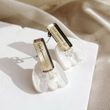 A pair of elegant earrings featuring rectangular gold elements and white, iridescent, shell-like shapes, designed for allergy-free comfort, displayed on a white surface with a soft, beige fabric background.