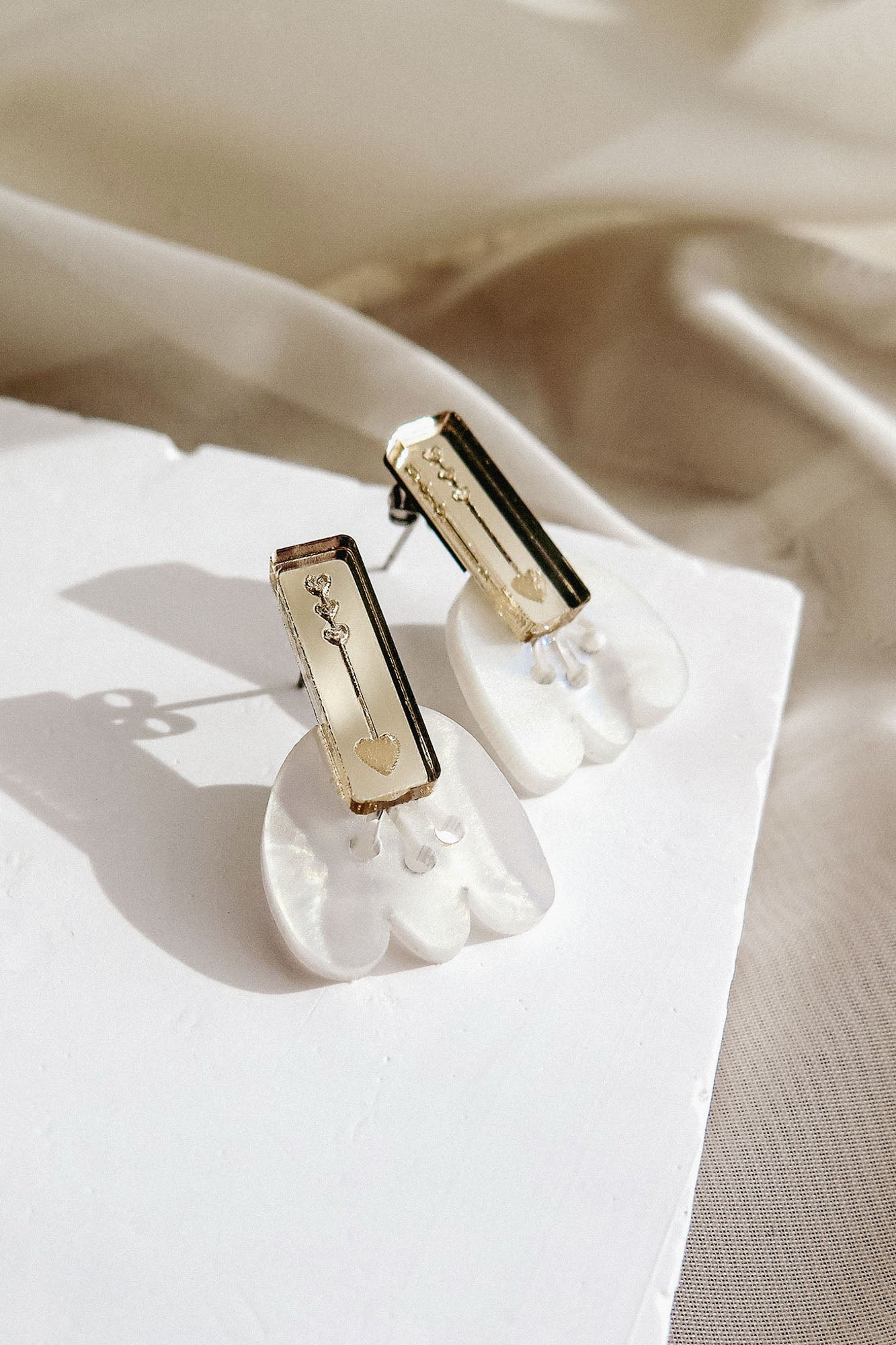 A pair of elegant earrings featuring rectangular gold elements and white, iridescent, shell-like shapes, designed for allergy-free comfort, displayed on a white surface with a soft, beige fabric background.