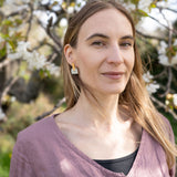 A woman with long blonde hair, wearing a mauve top and Romantic Tulip Earrings in marbled purple acrylic, stands outdoors in front of a blooming tree with white flowers. Sunlight highlights her face and the spring blossoms behind her.