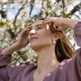 A woman wearing a mauve blouse and Romantic Tulip Earrings stands outside with her hands near her face, eyes closed, enjoying sunlight beneath blossoming tree branches. The marbled purple acrylic adds an allergy-free comfort to her serene moment.